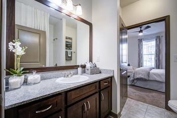 A bathroom with a large mirror, sink, and a bedroom in the background.